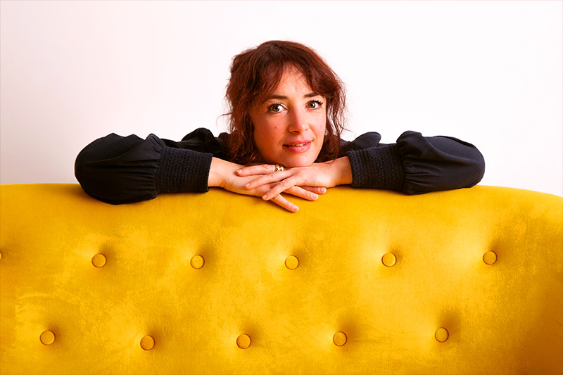 Portrait photo de Fanny VAURY, bras croisés sur le dossier d'un canapé jaune.